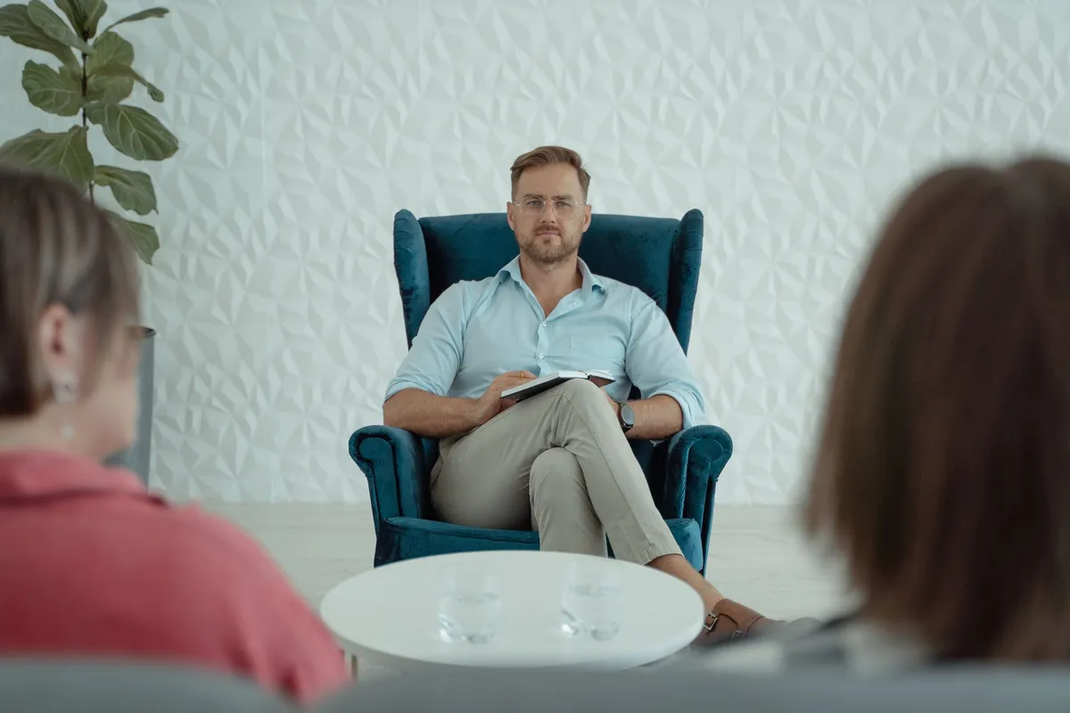 A counsellor sitting in an armchair with a notepad, facing a couple seen from behind