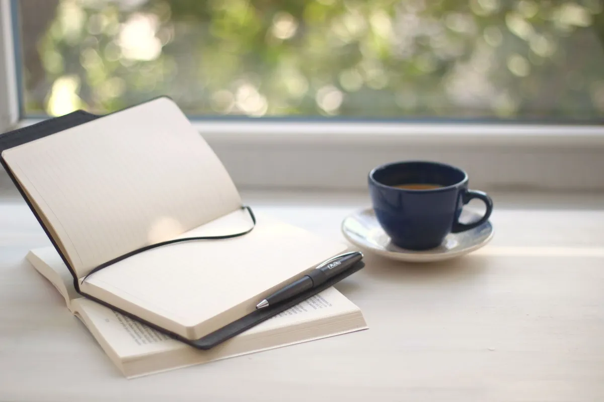 An open notebook and pen beside a cup of coffee on a windowsill in daylight