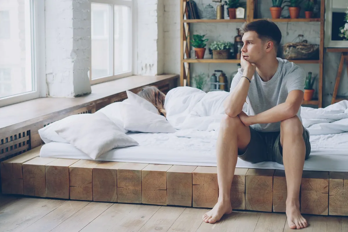 A man sitting on the edge of a bed in the morning looking away