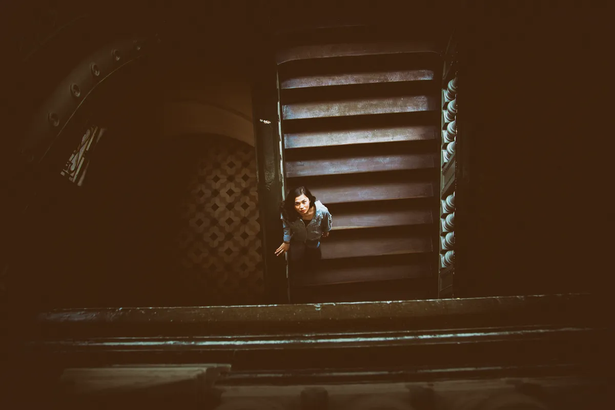 A woman standing at the bottom of a dark stairwell looking up towards the light