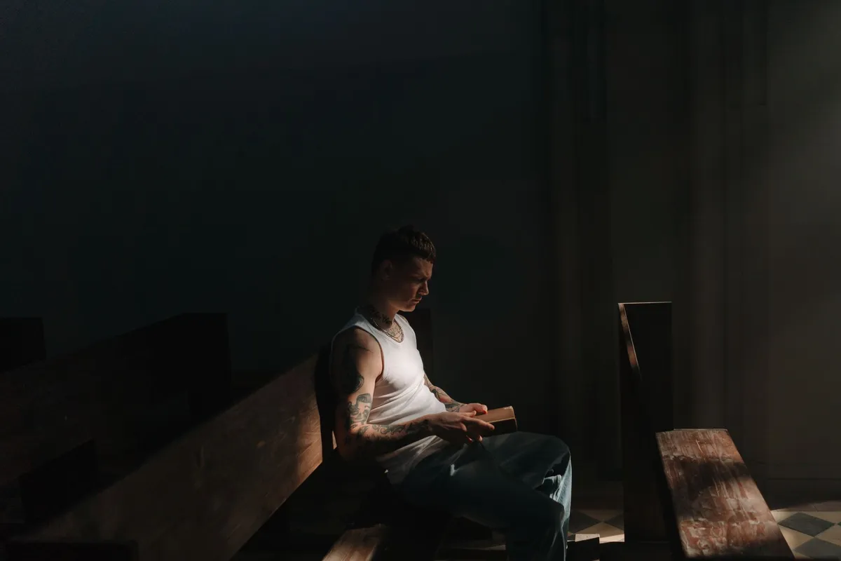 A man sitting alone in a dark church pew holding a book in a shaft of light