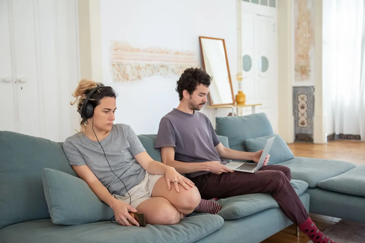 A couple sitting back to back on a sofa, one wearing headphones and the other on a laptop